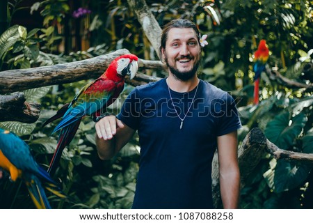  Young handsome man with a beard posing in the zoo he has on his hands a multicolored talking parrots, he smiles, macho, mustache, animal world, garden, smile, bird, Bali