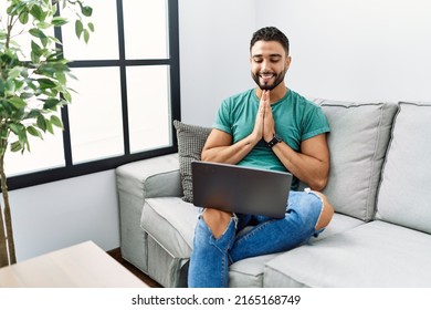 Young Handsome Man With Beard Using Computer Laptop Sitting On The Sofa At Home Praying With Hands Together Asking For Forgiveness Smiling Confident. 