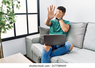 Young Handsome Man With Beard Using Computer Laptop Sitting On The Sofa At Home Covering Eyes With Hands And Doing Stop Gesture With Sad And Fear Expression. Embarrassed And Negative Concept. 