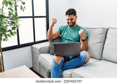 Young Handsome Man With Beard Using Computer Laptop Sitting On The Sofa At Home Very Happy And Excited Doing Winner Gesture With Arms Raised, Smiling And Screaming For Success. Celebration Concept. 