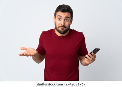 Young handsome man with beard using mobile phone isolated on white background making doubts gesture while lifting the shoulders