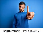 Young handsome man with beard wearing casual sweater and glasses over blue background Pointing with finger up and angry expression, showing no gesture