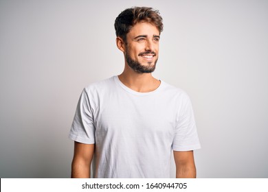 Young handsome man with beard wearing casual t-shirt standing over white background looking away to side with smile on face, natural expression. Laughing confident.