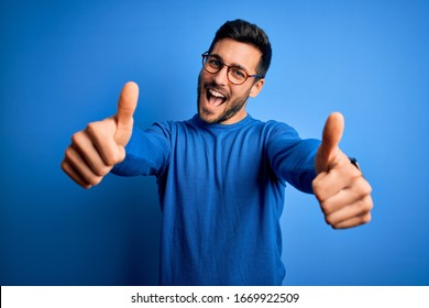 Young handsome man with beard wearing casual sweater and glasses over blue background approving doing positive gesture with hand, thumbs up smiling and happy for success. Winner gesture.