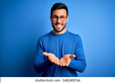Young handsome man with beard wearing casual sweater and glasses over blue background Smiling with hands palms together receiving or giving gesture. Hold and protection - Shutterstock ID 1682400766