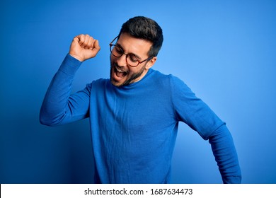 Young handsome man with beard wearing casual sweater and glasses over blue background Dancing happy and cheerful, smiling moving casual and confident listening to music