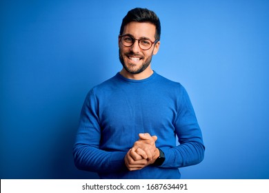 Young handsome man with beard wearing casual sweater and glasses over blue background with hands together and crossed fingers smiling relaxed and cheerful. Success and optimistic - Shutterstock ID 1687634491