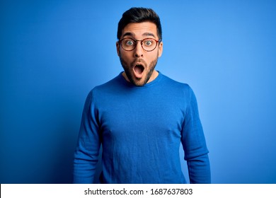 Young handsome man with beard wearing casual sweater and glasses over blue background afraid and shocked with surprise and amazed expression, fear and excited face. - Shutterstock ID 1687637803