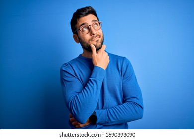 Young handsome man with beard wearing casual sweater and glasses over blue background Thinking worried about a question, concerned and nervous with hand on chin