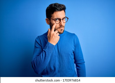 Young handsome man with beard wearing casual sweater and glasses over blue background Pointing to the eye watching you gesture, suspicious expression