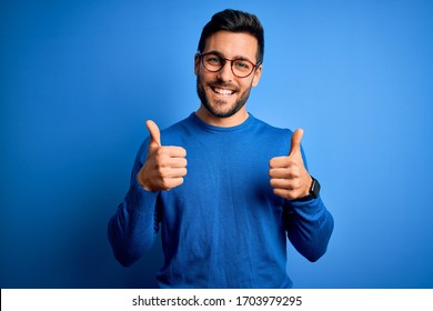 Young handsome man with beard wearing casual sweater and glasses over blue background success sign doing positive gesture with hand, thumbs up smiling and happy. Cheerful expression and winner  - Shutterstock ID 1703979295