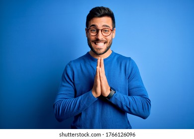 Young handsome man with beard wearing casual sweater and glasses over blue background praying with hands together asking for forgiveness smiling confident. - Shutterstock ID 1766126096