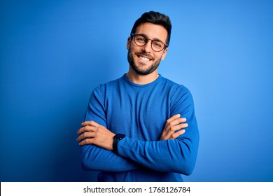 Junge gut aussehende Mann mit Bart, die einen ungezwungenen Pullover und eine Brille auf blauem Hintergrund trägt, glückliches Gesicht, das mit gekreuzten Armen die Kamera anschaut. Positive Person.