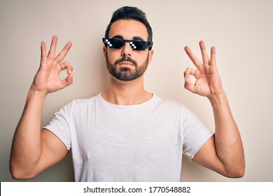 Young handsome man with beard wearing funny thug life sunglasses over white background relax and smiling with eyes closed doing meditation gesture with fingers. Yoga concept.