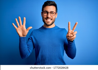 Young handsome man with beard wearing casual sweater and glasses over blue background showing and pointing up with fingers number seven while smiling confident and happy.
