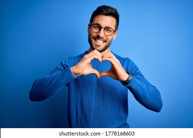 Young handsome man with beard wearing casual sweater and glasses over blue background smiling in love doing heart symbol shape with hands. Romantic concept.
