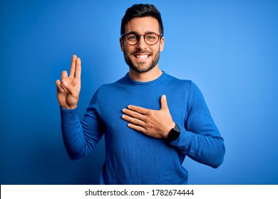 Young handsome man with beard wearing casual sweater and glasses over blue background smiling swearing with hand on chest and fingers up, making a loyalty promise oath
