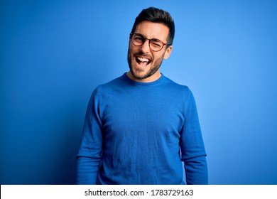Young handsome man with beard wearing casual sweater and glasses over blue background winking looking at the camera with sexy expression, cheerful and happy face. - Shutterstock ID 1783729163
