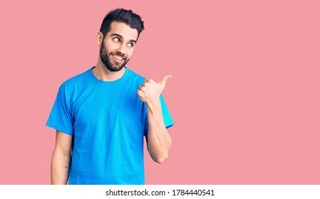 Young handsome man with beard wearing casual t-shirt smiling with happy face looking and pointing to the side with thumb up. 