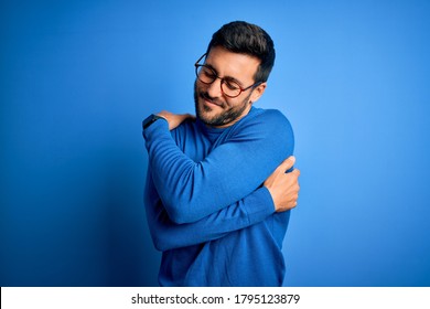 Young handsome man with beard wearing casual sweater and glasses over blue background Hugging oneself happy and positive, smiling confident. Self love and self care