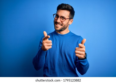 Young Handsome Man With Beard Wearing Casual Sweater And Glasses Over Blue Background Pointing Fingers To Camera With Happy And Funny Face. Good Energy And Vibes.