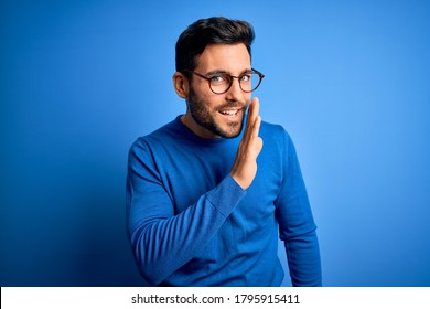 Young handsome man with beard wearing casual sweater and glasses over blue background hand on mouth telling secret rumor, whispering malicious talk conversation - Shutterstock ID 1795915411