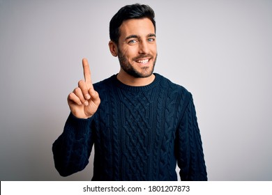 Young handsome man with beard wearing casual sweater standing over white background showing and pointing up with finger number one while smiling confident and happy.