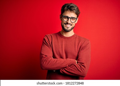 Young handsome man with beard wearing glasses and sweater standing over red background happy face smiling with crossed arms looking at the camera. Positive person.