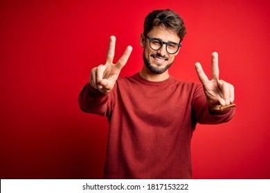 Young handsome man with beard wearing glasses and sweater standing over red background smiling with tongue out showing fingers of both hands doing victory sign. Number two.