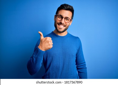 Young handsome man with beard wearing casual sweater and glasses over blue background doing happy thumbs up gesture with hand. Approving expression looking at the camera showing success.