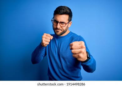 Young handsome man with beard wearing casual sweater and glasses over blue background Punching fist to fight, aggressive and angry attack, threat and violence