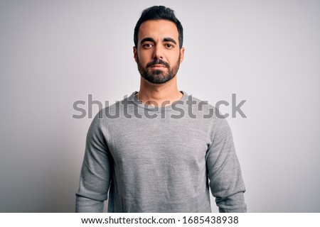 Young handsome man with beard wearing casual sweater standing over white background Relaxed with serious expression on face. Simple and natural looking at the camera.