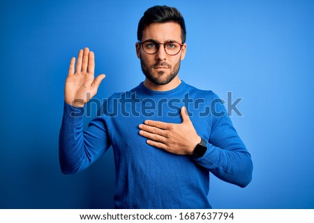 Young handsome man with beard wearing casual sweater and glasses over blue background Swearing with hand on chest and open palm, making a loyalty promise oath