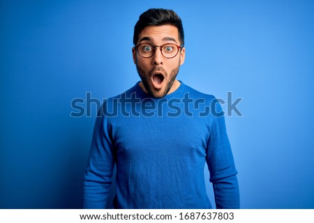 Young handsome man with beard wearing casual sweater and glasses over blue background afraid and shocked with surprise and amazed expression, fear and excited face.