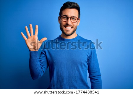 Young handsome man with beard wearing casual sweater and glasses over blue background showing and pointing up with fingers number five while smiling confident and happy.