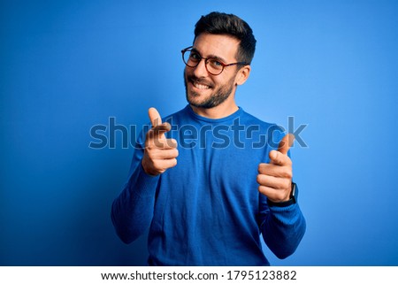 Young handsome man with beard wearing casual sweater and glasses over blue background pointing fingers to camera with happy and funny face. Good energy and vibes.