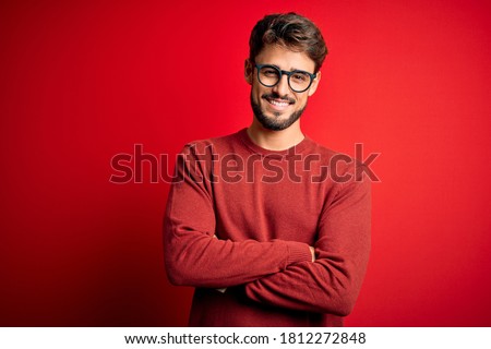 Young handsome man with beard wearing glasses and sweater standing over red background happy face smiling with crossed arms looking at the camera. Positive person.