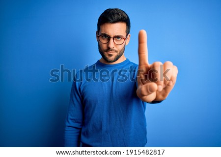 Young handsome man with beard wearing casual sweater and glasses over blue background Pointing with finger up and angry expression, showing no gesture
