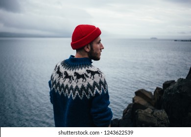 Young handsome man with beard wears traditional authentic blue wool knitted sweater with ornaments and red fisherman or sailor beanie hat, looks to horizon, pensative and thoughtful