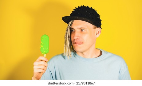 Young Handsome Man In Black Cap Looking At Ice Cream, Licking Lips On Yellow Background. Portrait Of Happy Male With Dreadlocks In Anticipation Enjoying Sundae