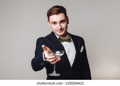 Young Handsome Man In Black Jacket And Bow Tie Holding A Drink And Making Toast Gesture Isolated On Grey Background. New Year's Eve. Offering A Drink