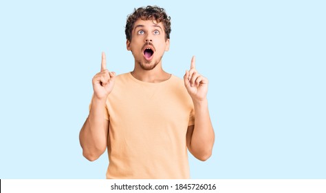 Young handsome man with curly hair wearing casual clothes amazed and surprised looking up and pointing with fingers and raised arms.  - Shutterstock ID 1845726016