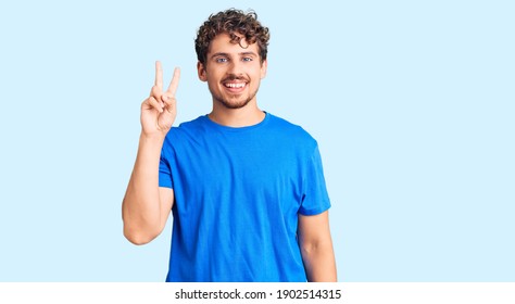 Young handsome man with curly hair wearing casual clothes showing and pointing up with fingers number two while smiling confident and happy. 