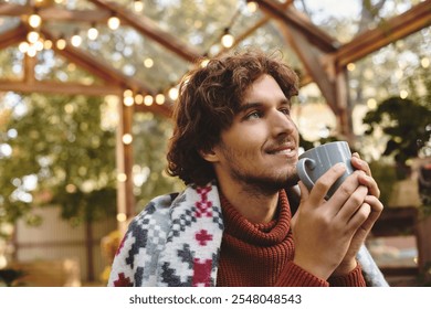 A young and handsome man with curly hair relaxes in a vibrant greenhouse, holding a steaming mug. Surrounded by lush greenery and fairy lights, he exudes warmth and tranquility. - Powered by Shutterstock