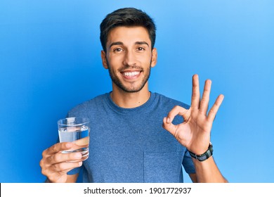 Young handsome man drinking glass of water doing ok sign with fingers, smiling friendly gesturing excellent symbol 