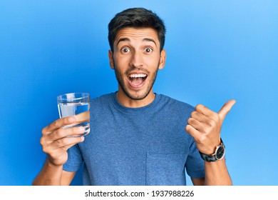 Young handsome man drinking glass of water pointing thumb up to the side smiling happy with open mouth 