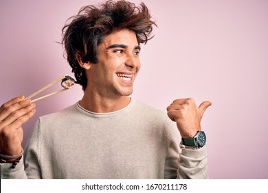 Young handsome man eating sushi using chopsticks over isolated pink background pointing and showing with thumb up to the side with happy face smiling