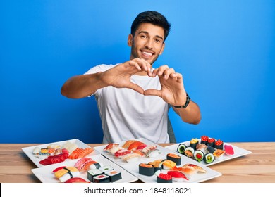 Young handsome man eating sushi sitting on the table smiling in love doing heart symbol shape with hands. romantic concept. 