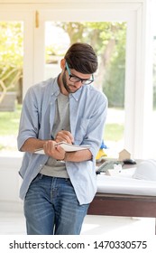 Young Handsome Man Engineer Working At Home. He Stand And Writing Report Near His Desk On His Holiday, Business Man Concept