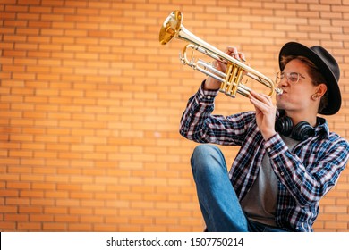 Young handsome man with eye glasses and hat poses with trumpet outdoor with brick wall